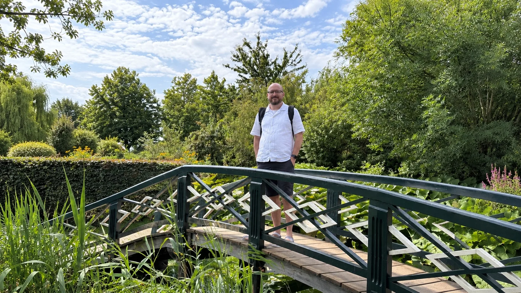 Brandon Seibel, founder of Trip7, standing on a green wooden footbridge in a garden, with a backpack, under a summer sky